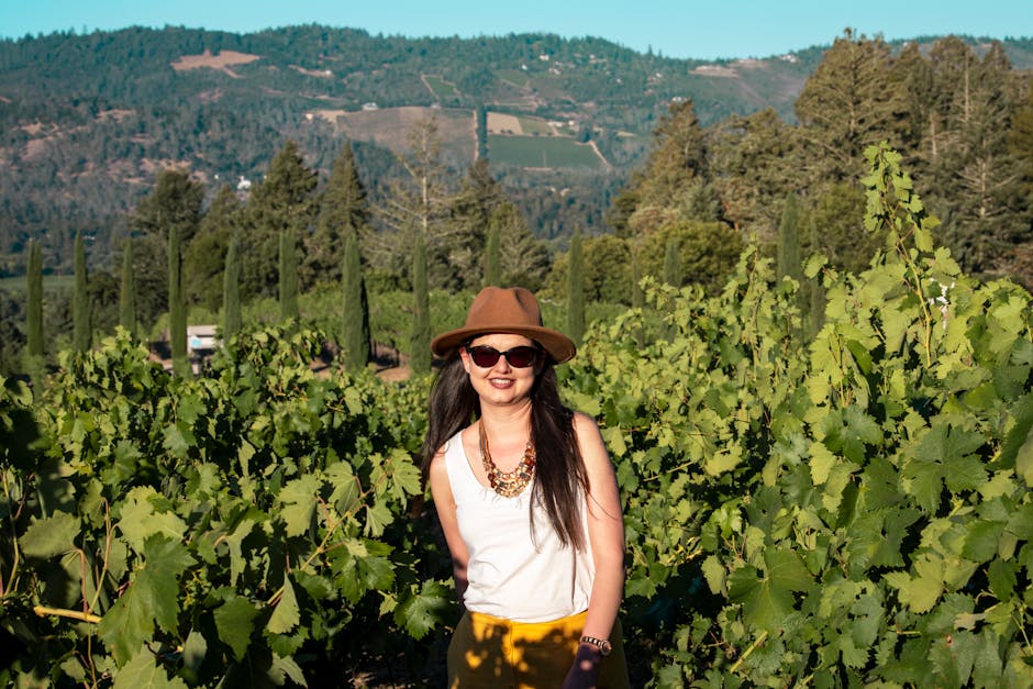 A woman in a hat and sunglasses smiles amidst lush vineyard with scenic mountain backdrop.